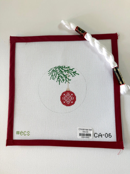 Cross-stitched design of a Christmas ball with a tree on fabric, with a needle and thread in the background.